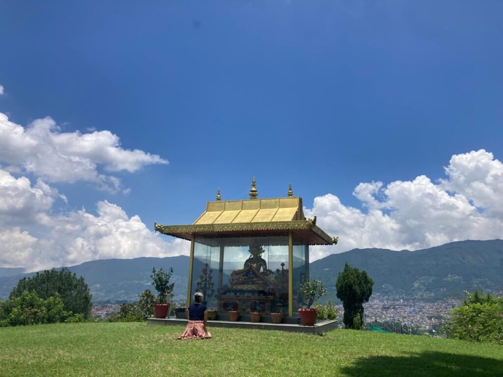 Mulher meditando em frente a uma estátua envidraçada no Kopan Monastery, com vista panorâmica do vale de Kathmandu, montanhas e céu azul